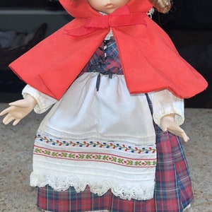 Puede incluir: Una muñeca de porcelana vestida con una capa roja con capucha, una blusa blanca y una falda a cuadros. La muñeca tiene el pelo rizado marrón y ojos azules.