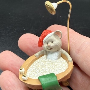 May include: A miniature figurine of a light brown mouse wearing a red and white Santa hat, sitting in a tan clawfoot bathtub filled with white bubble bath foam. A green towel hangs over the edge, and a gold shower head fixture stands above.
