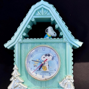 May include: A light blue Snoopy quartz clock in a house-shaped design. The clock face features Snoopy wearing sunglasses. A Snoopy figure is visible above the clock face. A Christmas tree and bell are on the sides.