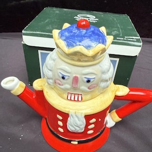 May include: A ceramic teapot shaped like a nutcracker king, featuring a red coat, yellow collar, and a blue crown with a red pom-pom. The figure has white hair, a pink nose, and a painted mouth. It is positioned in front of a green cardboard box.