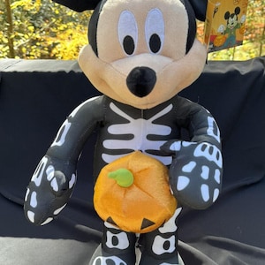 May include: A black and white skeleton Mickey Mouse plush toy holding a small orange pumpkin. The plush toy is wearing a black and white skeleton costume. The toy is standing on a black surface.