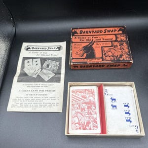Puede incluir: Un juego de mesa vintage llamado "Barnyard Swap" con una caja roja y negra y un juego de cartas. La caja presenta un burro de dibujos animados y el texto "A Game of Fun... For Old and Young!" Las cartas son rojas y blancas con ilustraciones de animales de granja.