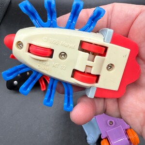 May include: A selection of vintage Marvel toy cars. The main car is off-white with red and blue details, blue appendages, and red wheels. The text "1995 Marvel" and "China MT 03" are visible.