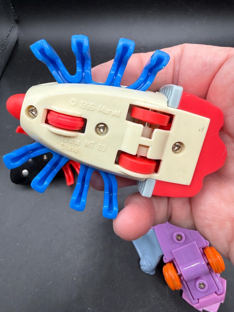 May include: A selection of vintage Marvel toy cars. The main car is off-white with red and blue details, blue appendages, and red wheels. The text "1995 Marvel" and "China MT 03" are visible.