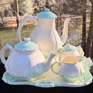 May include: A white and light blue ceramic tea set. The set includes a teapot, a creamer, a sugar bowl, and a serving tray. The teapot has a curved handle and a matching lid. The creamer and sugar bowl have a scalloped design.