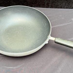 Könnte beinhalten: Eine schwarze Wok-Pfanne mit einem langen Griff und einem kleinen Schlaufengriff. Die Pfanne ist aus Metall und hat eine dunkle Innenseite. Der Griff ist schwarz und hat ein Loch am Ende zum Aufhängen. Ein Küchenutensil.