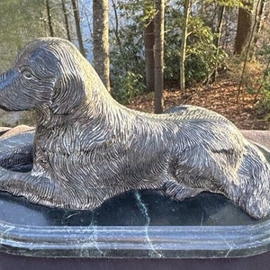 May include: A silver-colored sculpture of a dog lying down on a black marble base. The dog is depicted with realistic detail, including fur and facial features.