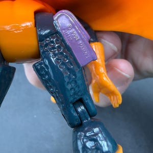 May include: Close-up of a vintage action figure's leg and arm. The figure has orange, dark blue, and purple parts. The purple piece is stamped with "&copy; 1994 MARVEL CHINA". The figure's arm is orange.