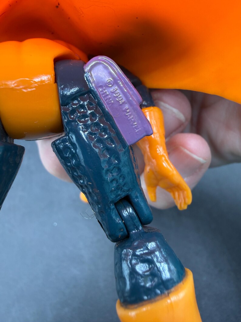 May include: Close-up of a vintage action figure's leg and arm. The figure has orange, dark blue, and purple parts. The purple piece is stamped with "&copy; 1994 MARVEL CHINA". The figure's arm is orange.