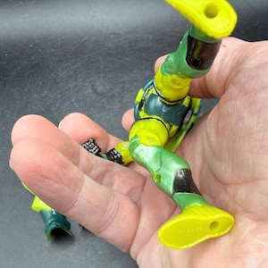 May include: A partially assembled action figure with a green and yellow color scheme. The figure's legs and torso are visible, with details including black accents and molded textures. The figure is held in a hand.