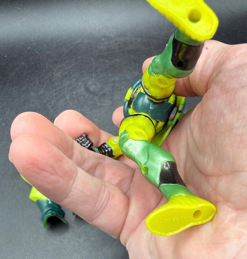 May include: A partially assembled action figure with a green and yellow color scheme. The figure's legs and torso are visible, with details including black accents and molded textures. The figure is held in a hand.