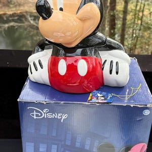 May include: A ceramic Mickey Mouse figure with a black head and ears, a tan face, and a red and white outfit. The figure is on top of a blue Disney box with Mickey and Minnie Mouse graphics.