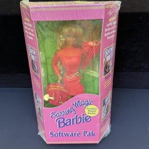Op de afbeelding: Een blonde Earring Magic Barbie-pop in een rode jurk met een gouden riem, verpakt in een roze doos met het label Earring Magic Barbie Software Pak. De doos bevat een rode ronde kaart met clip-oorbellen en tekst Genuine Mattel Barbie.