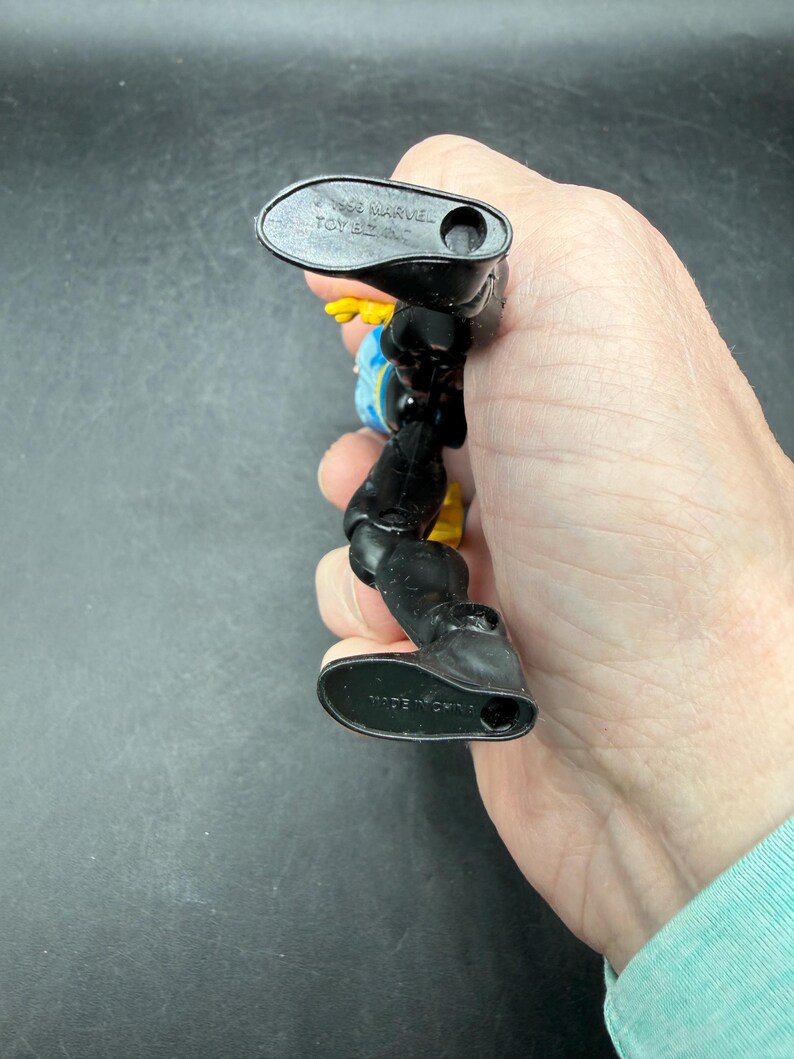 May include: A black and yellow action figure with blue accents. The figure is held in a hand, with the feet of the figure facing the camera. The feet have the text "MADE IN CHINA". The figure is marked "&copy; 1992 MARVEL TOY BIZ".