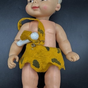 May include: A vintage plastic doll of a baby caveman wearing a brown and yellow animal skin loincloth and a bone necklace. The doll has white hair and a painted smile.
