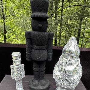 Może przedstawiać: Trzy dekoracyjne figurki: czarny żołnierz do dziadków do orzechów, mały srebrny dziadek do orzechów i srebrny krasnal ogrodowy. Czarny dziadek do orzechów jest najwyższy, z teksturowaną powierzchnią. Srebrne figurki mają lustrzany, odblaskowy wygląd.