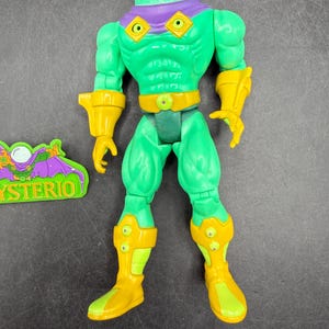 Op de afbeelding: Een groene actiefiguur van Mysterio, een stripboekschurk, met een heldere, ronde helm. De figuur heeft gele accenten op de handschoenen, laarzen en riem. Een apart groen en paars embleem met het woord "MYSTERIO" is ook aanwezig.