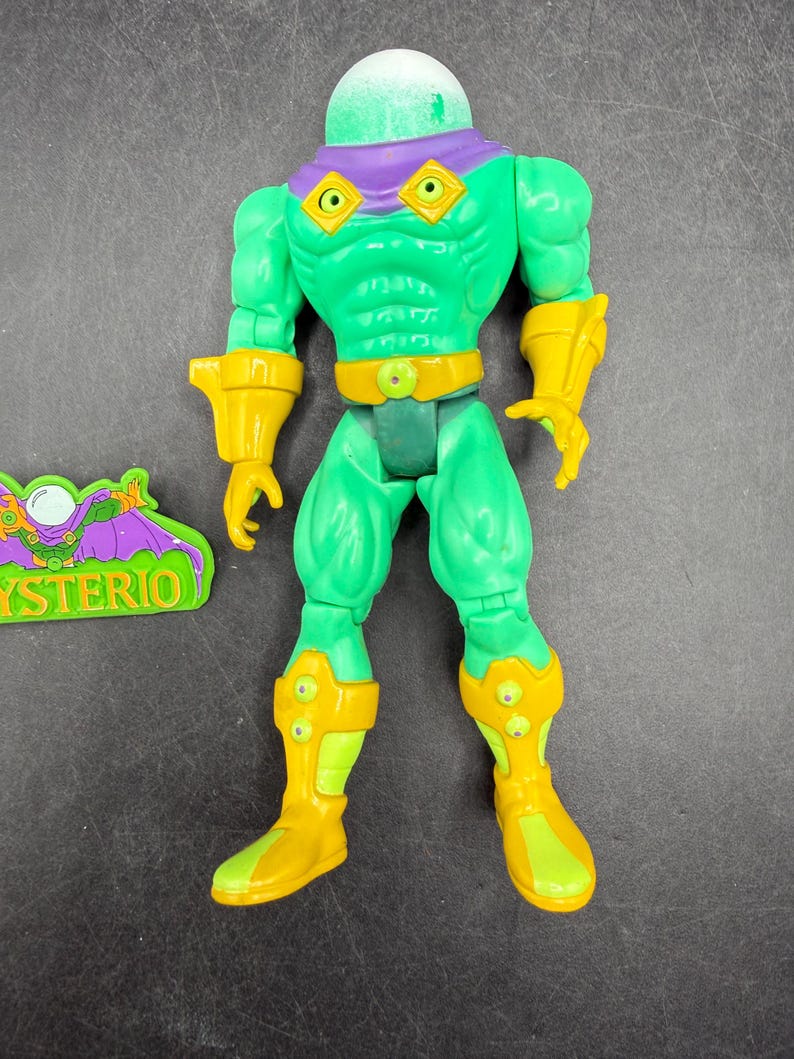 Op de afbeelding: Een groene actiefiguur van Mysterio, een stripboekschurk, met een heldere, ronde helm. De figuur heeft gele accenten op de handschoenen, laarzen en riem. Een apart groen en paars embleem met het woord "MYSTERIO" is ook aanwezig.