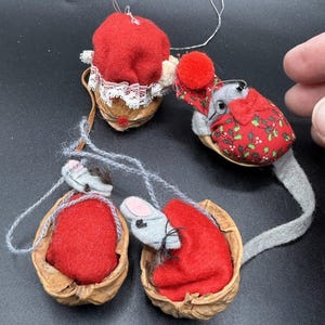 May include: Handmade Christmas ornaments featuring mice nestled in walnut shells. The mice are crafted from gray felt with red clothing and are adorned with small details. The ornaments are suspended by thin strings, perfect for holiday decor.