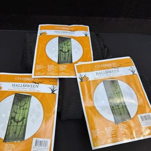 Op de afbeelding: Drie pakketten Halloween-raamgordijnen. Elk pakket toont een ontwerp van vleermuizen en spinnen tegen een maanverlichte achtergrond. De verpakking is oranje met de woorden "Celebrate It" en "Halloween". De gordijnen meten 101 cm x 213 cm.