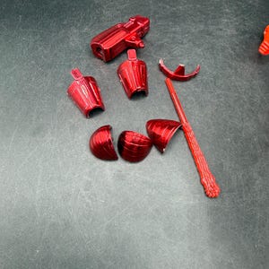 Puede incluir: Un juguete de pl&aacute;stico rojo desmontado, con varios componentes como una pieza en forma de pistola, secciones tipo armadura y una varilla larga y texturizada. Las piezas est&aacute;n dispersas sobre una superficie oscura.