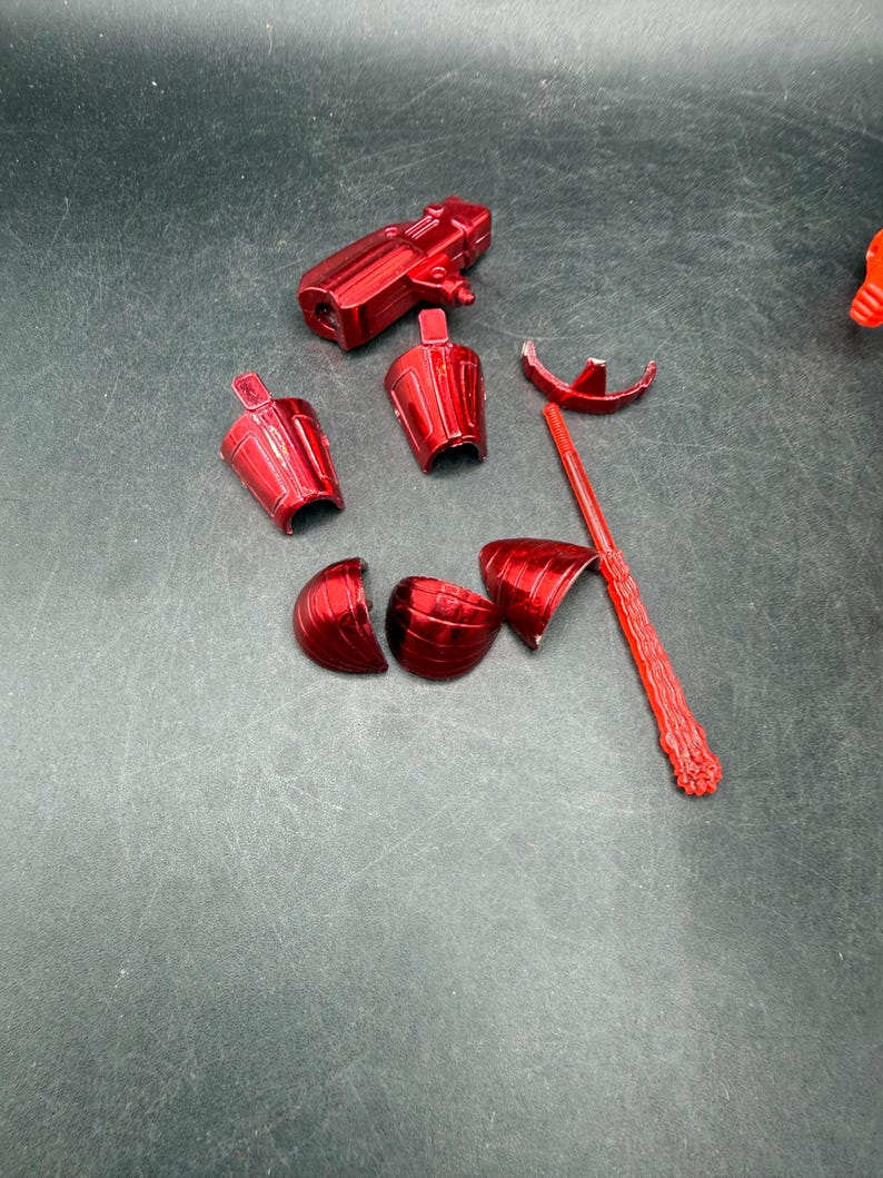 Puede incluir: Un juguete de pl&aacute;stico rojo desmontado, con varios componentes como una pieza en forma de pistola, secciones tipo armadura y una varilla larga y texturizada. Las piezas est&aacute;n dispersas sobre una superficie oscura.