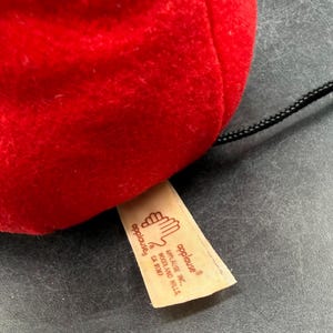May include: Close-up of a red plush toy with a black string attached. A tan tag with the "applause" logo and company information is visible. The toy is made of a soft, fuzzy material.