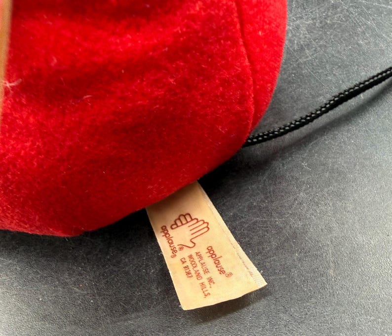 May include: Close-up of a red plush toy with a black string attached. A tan tag with the "applause" logo and company information is visible. The toy is made of a soft, fuzzy material.