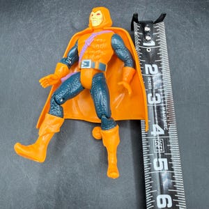 May include: An orange and blue action figure with a hooded head and cape. The figure is approximately 5 inches tall, with orange boots and gloves. The figure has a belt and a purple strap across the chest.
