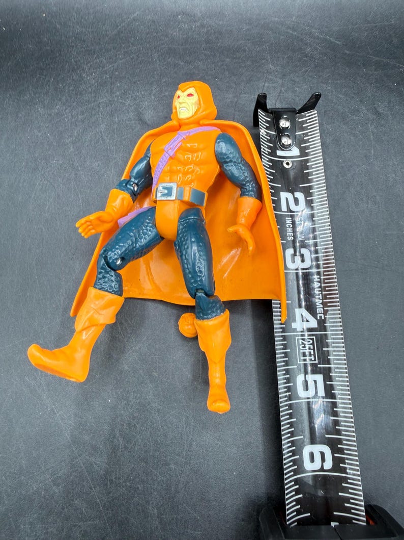 May include: An orange and blue action figure with a hooded head and cape. The figure is approximately 5 inches tall, with orange boots and gloves. The figure has a belt and a purple strap across the chest.