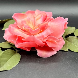 May include: A large, open pink rose with multiple layers of petals, accented with yellow hues in the center. The rose is surrounded by several green leaves, all set against a dark background. This floral arrangement is ideal for home decor.