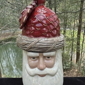 May include: Ceramic Santa Claus cookie jar with a red and brown pine cone lid. The jar features a white beard and a smiling face.