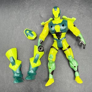 May include: A yellow and green action figure with detachable armor pieces. The figure has a helmet, chest armor, and leg armor. The figure is posed with one arm raised and the other holding a piece of armor. The figure is on a gray surface.