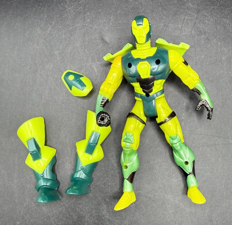 May include: A yellow and green action figure with detachable armor pieces. The figure has a helmet, chest armor, and leg armor. The figure is posed with one arm raised and the other holding a piece of armor. The figure is on a gray surface.