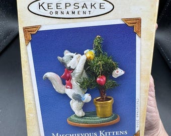 Könnte beinhalten: Hallmark Keepsake Ornament-Box mit einer grau-weißen Katze mit roter Schleife, die nach Ornamenten an einem kleinen Weihnachtsbaum greift. Auf der Box steht "Mischievous Kittens" und "Collector's Series".