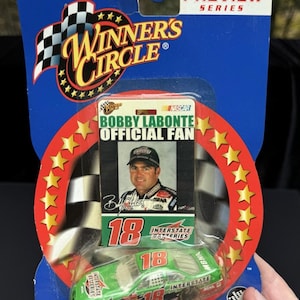 May include: A NASCAR Winners Circle Bobby Labonte Official Fan die-cast race car toy. The green and white car has the number 18 on it, and is packaged with a card featuring Bobby Labonte's photo. The packaging includes the text "PIT PASS PREVIEW SERIES".