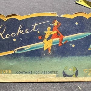 Puede incluir: Tarjeta vintage que anuncia agujas de coser de la marca Rocket. La tarjeta presenta un fondo azul con estrellas y un planeta. Dos figuras están montadas en una aguja con forma de cohete. El texto de la tarjeta dice "Rocket", "Silver", "Contiene 100 Surtidas", y "Gold Eye".