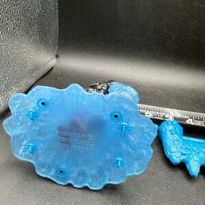 May include: A blue plastic toy base with a textured design and several holes. The base has the text "&copy; 1995 MARVEL TOY BIZ, INC. MADE IN CHINA". A blue plastic toy is next to the base.