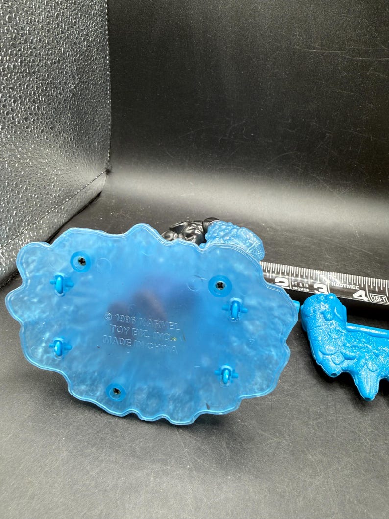 May include: A blue plastic toy base with a textured design and several holes. The base has the text "&copy; 1995 MARVEL TOY BIZ, INC. MADE IN CHINA". A blue plastic toy is next to the base.