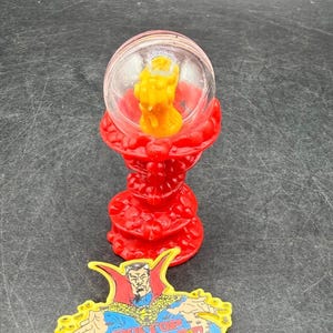 K&ouml;nnte beinhalten: Ein roter, verzierter St&auml;nder tr&auml;gt eine klare Kugel mit einer gelben Figur. Darunter befindet sich eine farbenfrohe Anstecknadel mit Doctor Strange und dem Text "DOCTOR STRANGE". Die Artikel sind wahrscheinlich Sammlerst&uuml;cke oder Spielzeug.