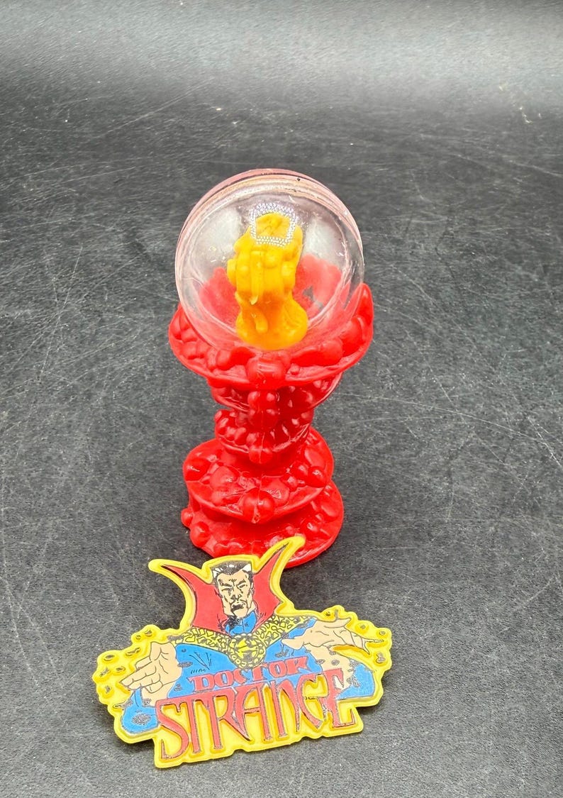 K&ouml;nnte beinhalten: Ein roter, verzierter St&auml;nder tr&auml;gt eine klare Kugel mit einer gelben Figur. Darunter befindet sich eine farbenfrohe Anstecknadel mit Doctor Strange und dem Text "DOCTOR STRANGE". Die Artikel sind wahrscheinlich Sammlerst&uuml;cke oder Spielzeug.