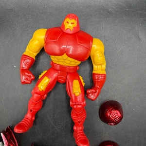 May include: A red and yellow Iron Man action figure with detachable parts. The figure has a red helmet, chest, and legs, with yellow arms and accents. Several red and yellow accessories are scattered around the figure.