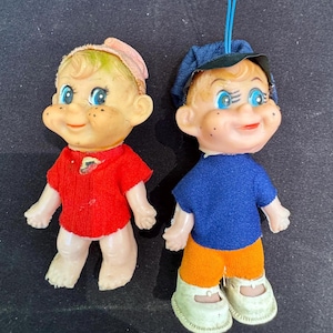 May include: Two vintage doll figures with smiling faces, blue eyes, and freckles. One doll wears a red shirt and a pink cap. The other doll is dressed in a blue shirt, orange pants, and a blue cap. Both dolls have white shoes.