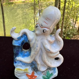 May include: A ceramic octopus figurine in shades of white, blue, and red. The octopus has eight tentacles and is perched on a blue and white base with a starfish and seaweed. The figurine is a decorative item.