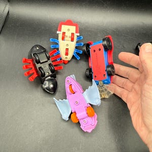 May include: Four colourful toy vehicles, including a red and blue car, a black bug, a pink bat, and a white bug. The toys have wheels and are made of plastic. The toys are on a black surface.