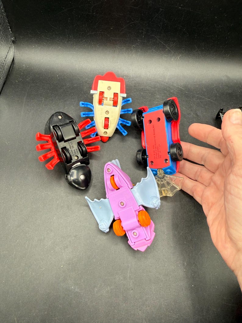 May include: Four colourful toy vehicles, including a red and blue car, a black bug, a pink bat, and a white bug. The toys have wheels and are made of plastic. The toys are on a black surface.