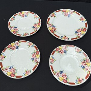 Puede incluir: Cuatro platos de cerámica blanca con borde rojo y diseño floral de frutas. Los platos presentan un patrón de rosas rosas, limones amarillos, manzanas rojas y bayas azules. Los platos están dispuestos sobre una superficie negra.