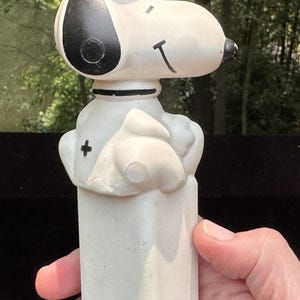 Può includere: Un distributore PEZ vintage di Snoopy. Il distributore presenta la testa di Snoopy con orecchie nere, un occhio nero e un cappello rosso. Il distributore è bianco con una base rettangolare ed è tenuto da una mano.