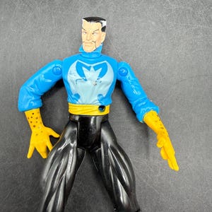 May include: A plastic action figure of a character with a stern expression. The figure wears a blue top with a light blue emblem, black pants, yellow gloves, and a yellow belt. The figure is posed against a gray background.