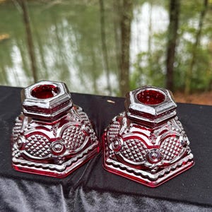 Könnte beinhalten: Zwei rote Glas-Kerzenhalter mit geometrischem Design. Die Kerzenhalter haben eine quadratische Basis und eine sechseckige Oberseite. Das Glas hat ein strukturiertes Muster mit Rautenformen und kreisförmigen Akzenten. Die Kerzenhalter werden auf einer schwarzen Oberfläche präsentiert.